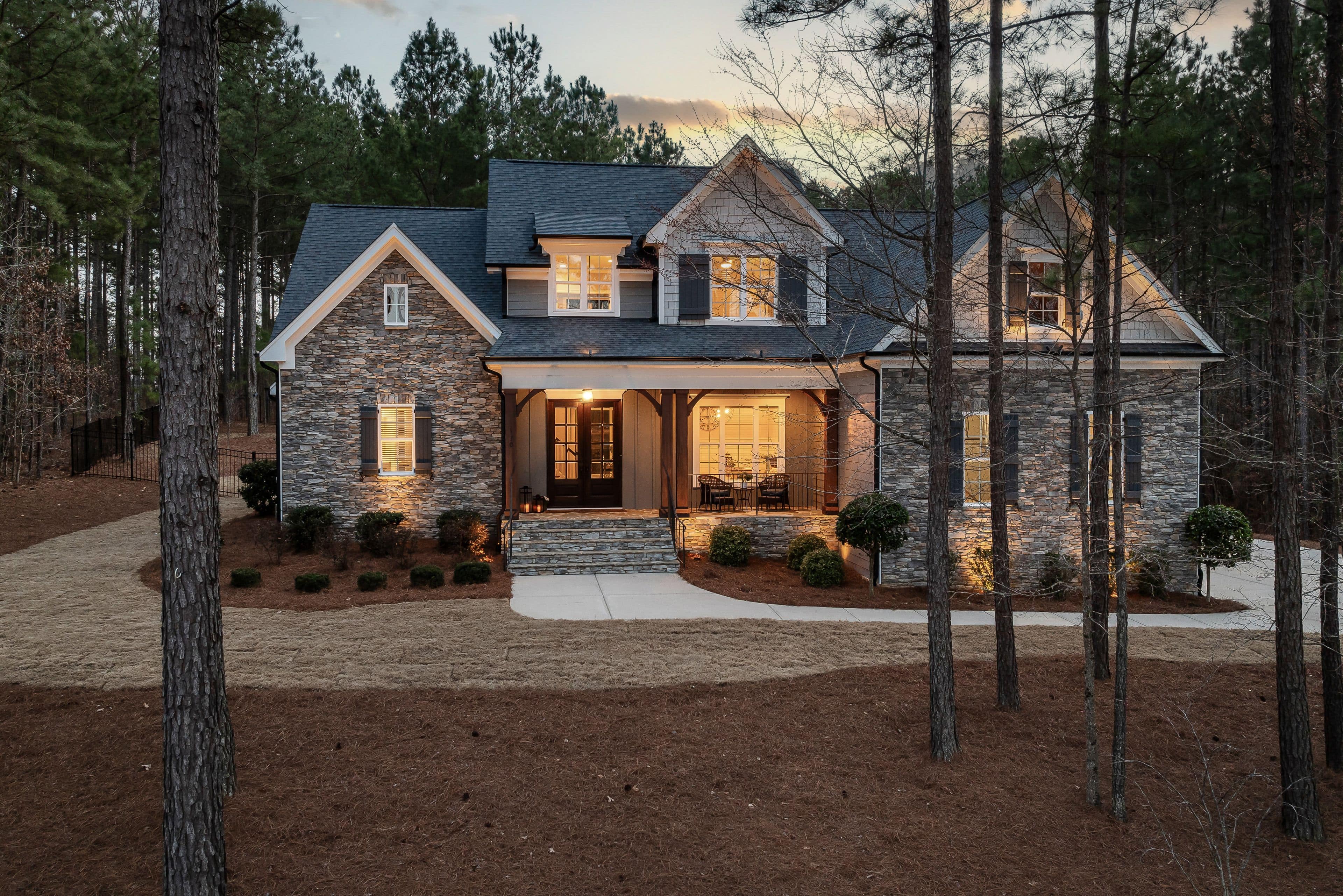 A stone-and-shingle family house at dusk, every window lit, seen through tall pines.