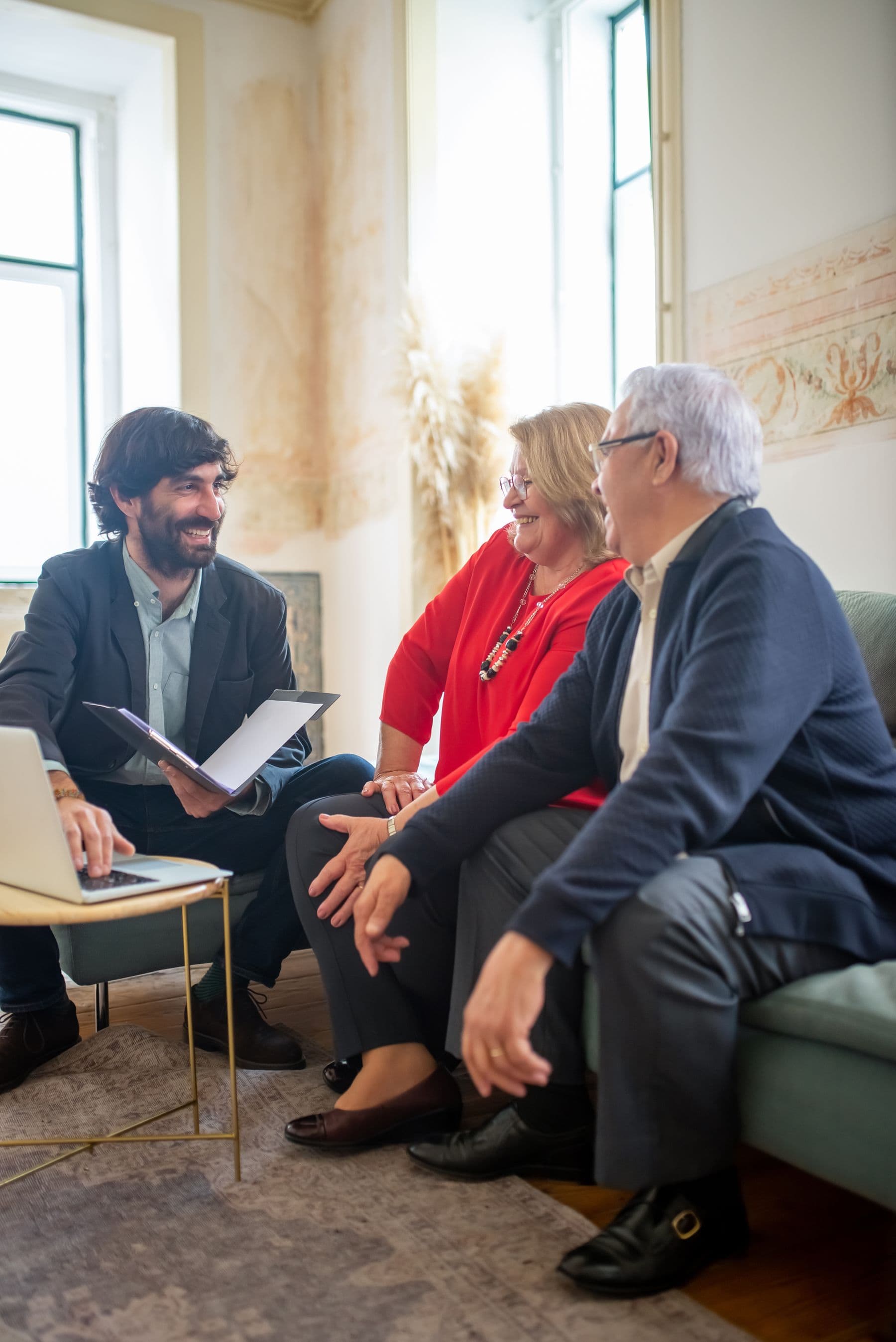 A man with an open folder and a laptop sits talking with an older couple on a sofa in a sunlit room.