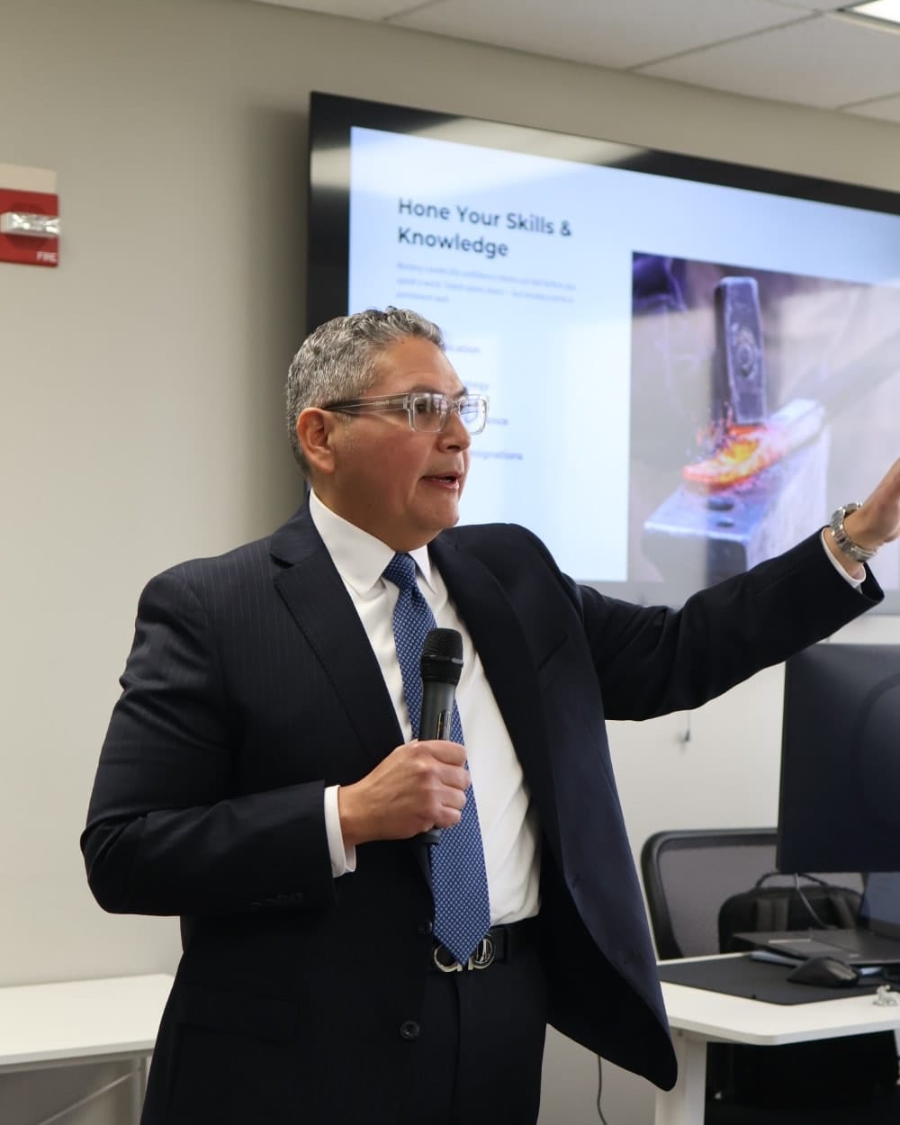 A Synergy advisor speaking into a microphone, gesturing towards a presentation slide headed “Hone Your Skills & Knowledge”.