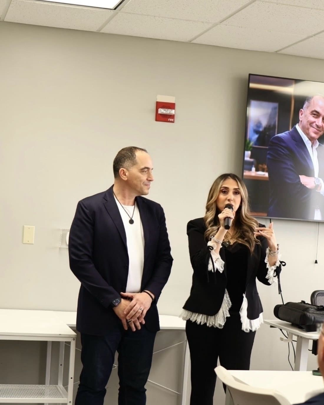 Two members of the Synergy team standing together at the front of a training room, one speaking into a microphone.